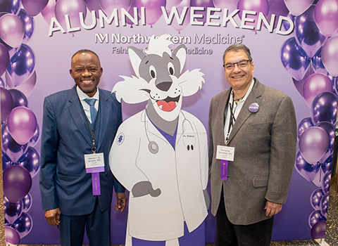 Two men standing with a cardboard cutout of Dr. Willie Wildcat in front of an Alumni Weekend step and repeat background