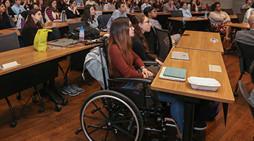 Trainees, including one in a wheel chair, sit at a table listening to a presentation.