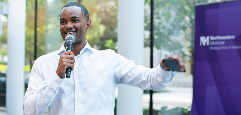 A speaker addresses the audience, holding a microphone and phone in hand