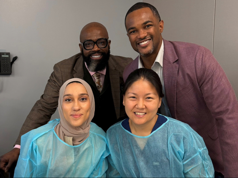 Two residents in blue scrubs and two men in suits, pose for a group image