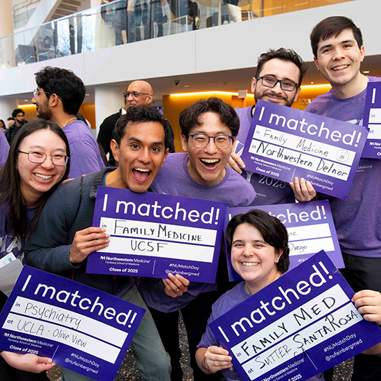 A group of medical school graduates smile and pose with signs showing where they will complete their residencies.