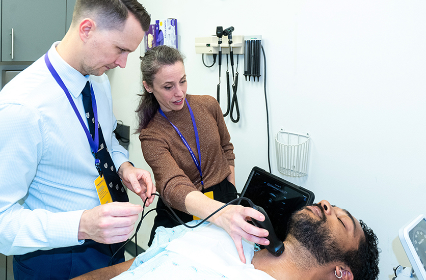 A medical educator at the Center for Bedside Medicine demonstrating the use of a handheld point-of-care ultrasound (POCUS) device on a patient while a physician participant observes the tablet interface.