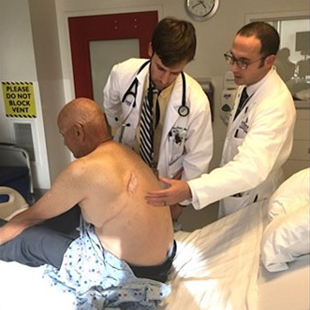 A clinical faculty preceptor observing a physician performing a physical examination on a patient's back to evaluate bedside clinical skills as part of the APECS program.