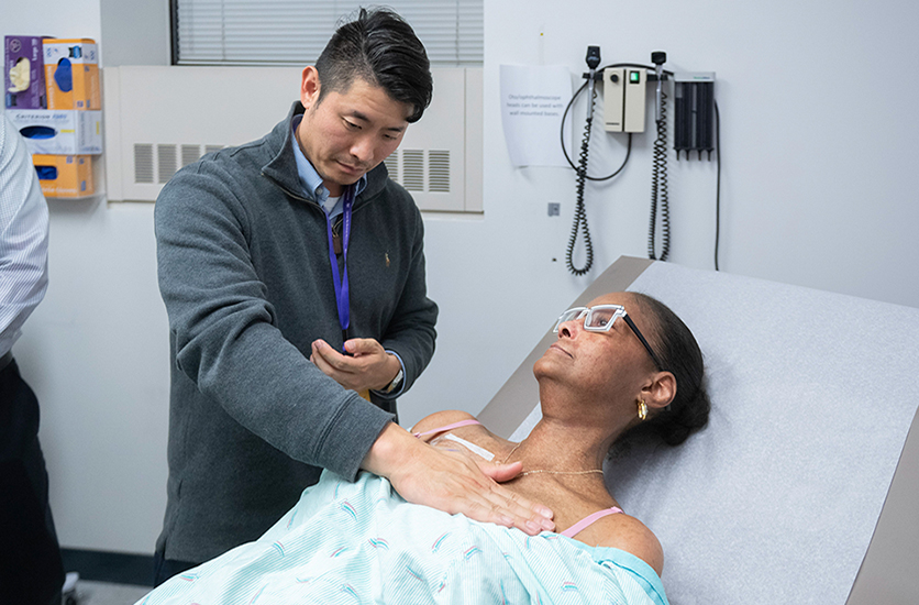 A physician participant in the APECS program performing a manual physical examination on a patient’s midsection while being observed by a clinical faculty preceptor in a simulation room.