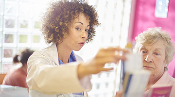 A middle aged woman highlights a brochure to an older woman in a clinical setting.