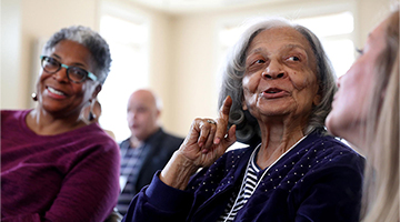An older woman appears to be talking, animating her point with by holding up her index finger. Other older adults are seated in the background looking happily engaged.