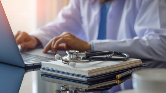 A doctor typing on a laptop with a stethoscope resting on a notebook nearby.