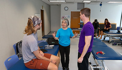 An instructor talks to two learners in a room of mat tables